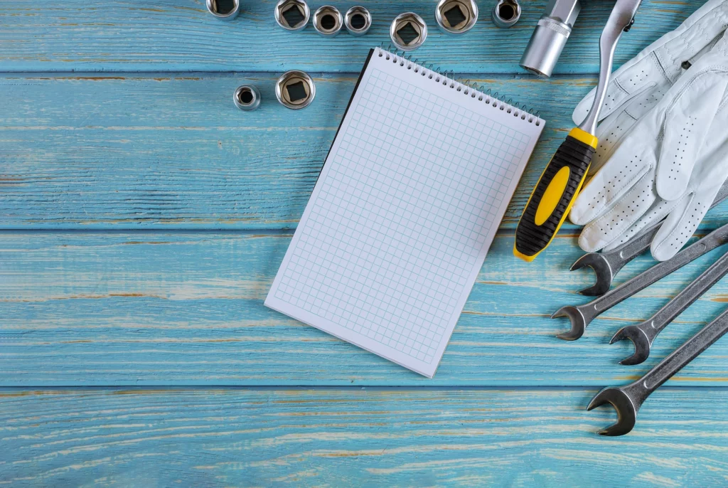 Arbeitshandschuhe, Schraubenschlüssel, Nüsse und ein Notizblick auf Holzuntergrund